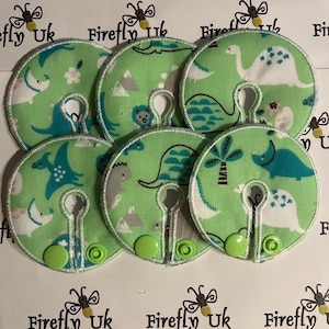 May include: Six green fabric nipple covers with a dinosaur print and a white border. The covers have a hole in the centre and a green button on the side. The covers are arranged in a circle on a white surface. The text "Firefly Uk" is printed on the surface.