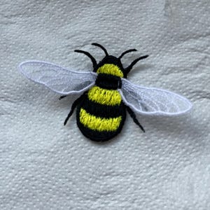May include: Embroidered bee patch with a black and yellow striped body, black antennae, and white wings. Ideal for sewing or ironing onto fabric. This patch is a decorative element for clothing or accessories.