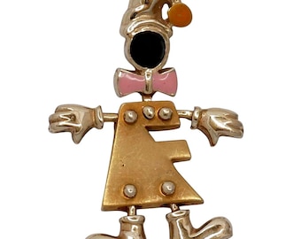 May include: Gold-toned pendant necklace featuring a whimsical figure. The figure has a black face, pink bow tie, and a hat with an orange accent. The body is a stylized shape with silver-colored details, and the arms and legs are outstretched.