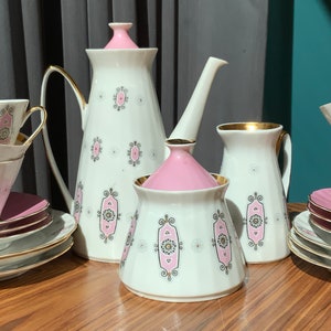 K&ouml;nnte beinhalten: Vintage Teeservice mit wei&szlig;em Grund, rosa Akzenten und goldenen Verzierungen. Das Set beinhaltet eine Teekanne, einen Sahnek&auml;nnchen, eine Zuckerdose, Tassen und Untertassen. Jedes Teil hat ein sich wiederholendes rosa-goldenes geometrisches Design.