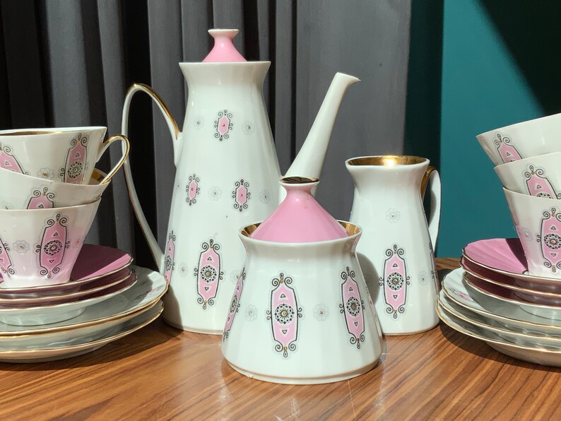 K&ouml;nnte beinhalten: Vintage Teeservice mit wei&szlig;em Grund, rosa Akzenten und goldenen Verzierungen. Das Set beinhaltet eine Teekanne, einen Sahnek&auml;nnchen, eine Zuckerdose, Tassen und Untertassen. Jedes Teil hat ein sich wiederholendes rosa-goldenes geometrisches Design.