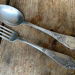 May include: A silver fork and spoon with ornate handles. The spoon features a floral design and the fork has a fan-shaped design. Both pieces have a monogrammed initial "H" on the back.