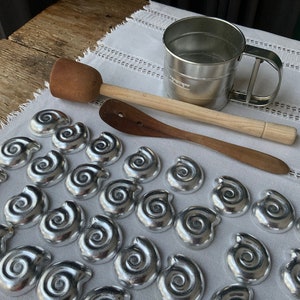 Op de afbeelding: Een assortiment keukengerei en decoratieve items ligt op een witte doek. De items omvatten een metalen bloemzeef, een houten stamper, een houten lepel en talrijke zilverkleurige, spiraalvormige decoratieve stukken.