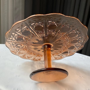 May include: A vintage amber-colored glass cake stand with a scalloped edge. The stand features a detailed geometric pattern on the plate and a pedestal base. The cake stand is designed to elevate and display baked goods.