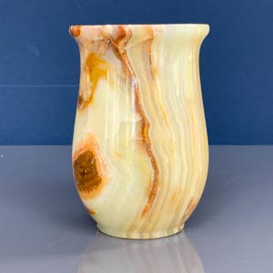 May include: An onyx vase with a flared top. The vase displays a mix of creamy white, yellow, and brown hues, with natural veining patterns. The smooth, polished surface reflects light, highlighting the stone's unique characteristics.