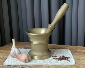 May include: A brass mortar and pestle with a wooden handle, two cloves of garlic, and a pile of cloves on a white linen placemat.