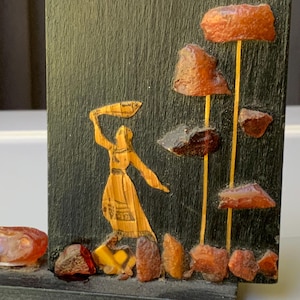 Peut inclure: Panneau décoratif avec une figure dorée sculptée dans une robe fluide, tenant un tissu. La figure est placée sur un fond sombre. Des pierres de couleur ambre sont disposées à la base et le long de lignes verticales.