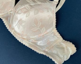 May include: Ivory-colored bra with a floral pattern in light pink and white. The bra has adjustable straps and a scalloped edge along the bottom. The fabric has a satin-like sheen.