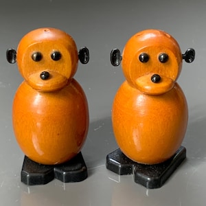 Puede incluir: Dos figuras de mono de madera con cuerpos naranjas, orejas negras y ojos negros. Los monos se apoyan sobre bases rectangulares negras. Las figuras tienen un acabado brillante y una textura suave.