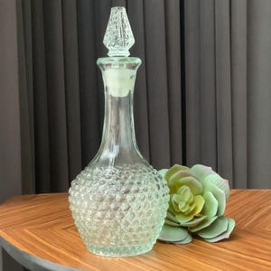 Może przedstawiać: Przezroczysta szklana karafka z teksturowaną podstawą w stylu hobnail i ozdobnym korkiem. Korek ma diamentowy wzór. Karafka jest eksponowana na drewnianej powierzchni z zielono-różowym sukulentem.