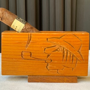 Op de afbeelding: Houten sigaren houder met een gesneden ontwerp van een man die een pijp rookt. De houder is gemaakt van lichtbruin hout en heeft een rechthoekige vorm. Een bruine sigaar ligt in de houder.
