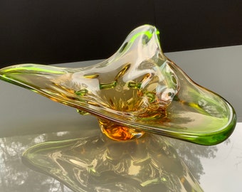 May include: A green and amber glass bowl with an abstract, organic shape. The bowl is made of blown glass and has a unique, flowing design.