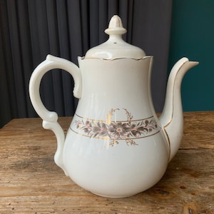 Puede incluir: Tetera de porcelana blanca con ribete dorado y diseño floral. La tetera tiene una tapa con un pomo en la parte superior.