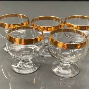 May include: Set of five clear glass drinking glasses with gold rims. The glasses have a round bowl and a short stem.