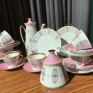 K&ouml;nnte beinhalten: Ein Vintage-Teeservice mit wei&szlig;em und rosa Blumendesign. Das Set umfasst Teetassen, Untertassen, Teller, eine Teekanne, einen Sahnek&auml;nnchen und eine Zuckerdose. Goldbesatz akzentuiert die R&auml;nder der Tassen und Teller. Das Set ist auf einem Holztisch angeordnet.