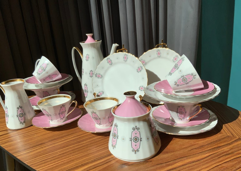 K&ouml;nnte beinhalten: Ein Vintage-Teeservice mit wei&szlig;em und rosa Blumendesign. Das Set umfasst Teetassen, Untertassen, Teller, eine Teekanne, einen Sahnek&auml;nnchen und eine Zuckerdose. Goldbesatz akzentuiert die R&auml;nder der Tassen und Teller. Das Set ist auf einem Holztisch angeordnet.