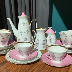 K&ouml;nnte beinhalten: Ein Vintage-Teeservice mit wei&szlig;er Porzellan-Teekanne, Sahnek&auml;nnchen, Zuckerdose, Tassen und Untertassen. Das Set hat ein rosa-goldenes Design mit einem dekorativen Muster. Die Tassen und Untertassen sind auf einer Holzoberfl&auml;che angeordnet.