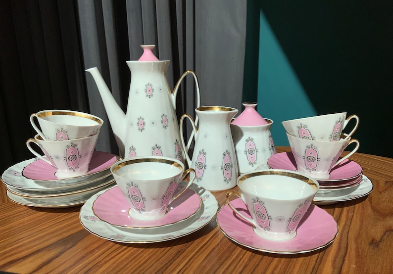K&ouml;nnte beinhalten: Ein Vintage-Teeservice mit wei&szlig;er Porzellan-Teekanne, Sahnek&auml;nnchen, Zuckerdose, Tassen und Untertassen. Das Set hat ein rosa-goldenes Design mit einem dekorativen Muster. Die Tassen und Untertassen sind auf einer Holzoberfl&auml;che angeordnet.