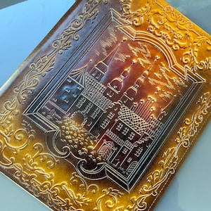 Puede incluir: Un diario encuadernado en cuero dorado con un diseño en relieve de un horizonte urbano. El diario tiene un borde decorativo con adornos de desplazamiento.