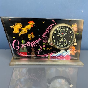 May include: A vintage clock with a black face featuring a rooster, the text "С добрым утром!" and a woman. The clock has a round clock face with three smaller dials. The clock is set on a clear base.