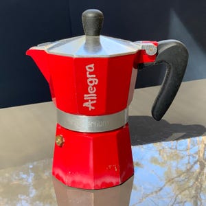 Puede incluir: Una cafetera italiana Allegra roja con asa negra y parte superior plateada. La cafetera está hecha de metal y tiene la palabra "Allegra" impresa en el lateral.