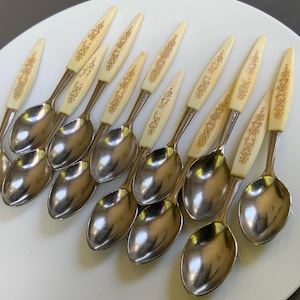 May include: A set of vintage spoons with cream-coloured handles and gold-coloured decorative patterns. The spoon bowls are silver. The spoons are arranged on a white surface.