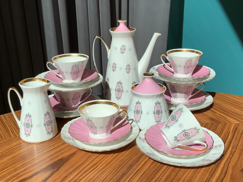 K&ouml;nnte beinhalten: Ein Vintage-Teeservice in Wei&szlig; und Rosa. Das Set umfasst eine Teekanne, einen Milchgie&szlig;er, eine Zuckerdose, Tassen und Untertassen. Jedes Teil ist mit Goldrand und einem sich wiederholenden rosa und grauen Muster verziert.