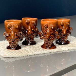 Può includere: Quattro bicchierini in ceramica marrone con un design a volto stravagante. Ogni bicchierino presenta un volto dettagliato con naso, occhi e barba prominenti. I bicchierini sono esposti su una superficie strutturata color crema.