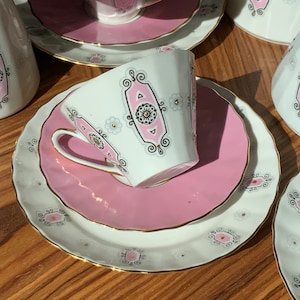 K&ouml;nnte beinhalten: Ein Vintage-Teeservice in Wei&szlig; und Rosa. Das Set umfasst Tassen, Untertassen und Servierteile, die mit einem rosa und schwarzen Design und goldenen Verzierungen verziert sind. Das Set ist auf einer Holzoberfl&auml;che angeordnet.