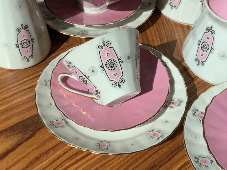 K&ouml;nnte beinhalten: Ein Vintage-Teeservice in Wei&szlig; und Rosa. Das Set umfasst Tassen, Untertassen und Servierteile, die mit einem rosa und schwarzen Design und goldenen Verzierungen verziert sind. Das Set ist auf einer Holzoberfl&auml;che angeordnet.