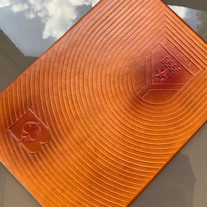 Puede incluir: Un diario encuadernado en cuero marrón con círculos concéntricos en relieve en la portada. El diario tiene una estrella roja y el texto "BAUSKA" en relieve en la portada.