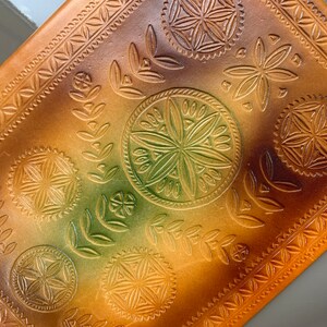 May include: A brown leather journal with a decorative embossed pattern. The pattern features a repeating design of leaves, flowers, and geometric shapes. The journal has a subtle ombre effect, transitioning from a darker brown to a lighter brown.