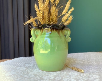 May include: A green ceramic vase with two handles and a brown rim. The vase is filled with dried wheat stalks.