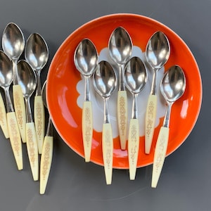 Op de afbeelding: Een set van tien zilveren theelepels met crèmekleurige handvatten met een gouden patroon. De lepels zijn gerangschikt op een fel oranje bord.