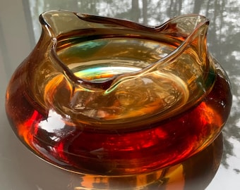 May include: A low-profile, wide-mouthed glass bowl featuring a deep amber and reddish-brown gradient. The rim has a distinct, undulating, wavy edge that creates a soft, organic silhouette. The glass is translucent, allowing light to pass through.