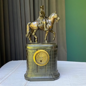 Puede incluir: Un reloj de bronce con una estatua de una persona a caballo en la parte superior. La esfera del reloj es redonda y tiene números romanos negros. El reloj está sobre una base negra con un patrón de ladrillo.