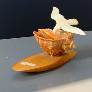 May include: A decorative ornament featuring a brown and orange conch shell mounted on an oval amber-coloured base. A cream-coloured bird figurine is attached to the top of the shell. The word "Capri" is written in white script on the base surface.