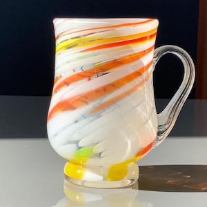 May include: A hand-blown glass mug with a clear handle and a colorful design. The mug features white, orange, yellow, and red swirls. The base of the mug is yellow. The mug is designed for hot beverages.