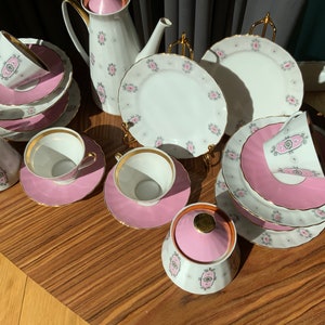 K&ouml;nnte beinhalten: Ein Vintage-Teeservice mit wei&szlig;er Basis, rosa Akzenten und goldenen Verzierungen. Das Set umfasst Tassen, Untertassen, Teller, einen Sahnek&auml;nnchen, eine Zuckerdose und eine Teekanne. Jedes St&uuml;ck hat ein rosa-graues Blumendesign.