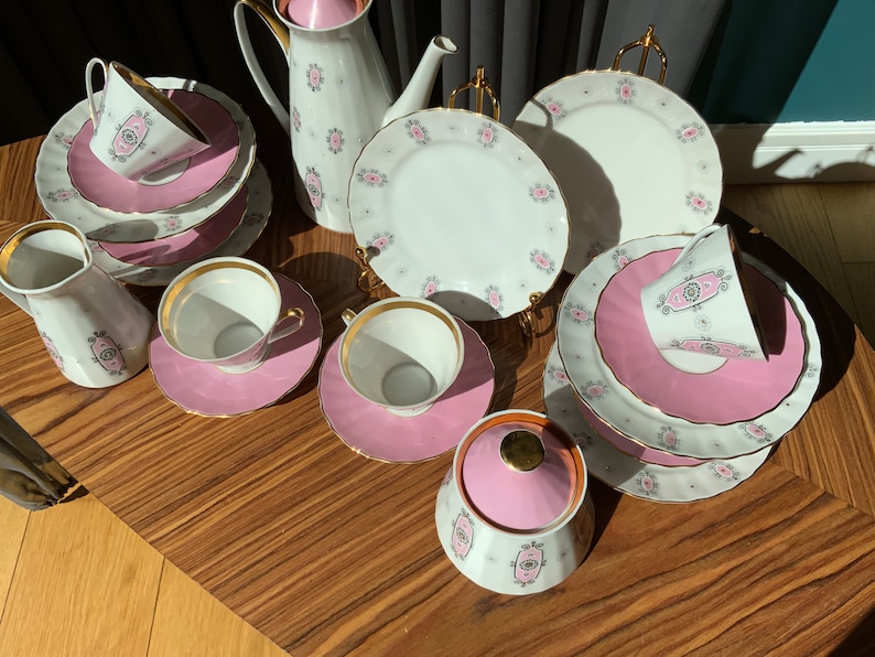 K&ouml;nnte beinhalten: Ein Vintage-Teeservice mit wei&szlig;er Basis, rosa Akzenten und goldenen Verzierungen. Das Set umfasst Tassen, Untertassen, Teller, einen Sahnek&auml;nnchen, eine Zuckerdose und eine Teekanne. Jedes St&uuml;ck hat ein rosa-graues Blumendesign.