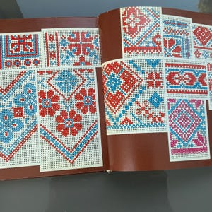 May include: An open book showing cross-stitch patterns in red and blue. The designs include geometric and floral motifs on a white background. The book is open on a brown surface.