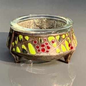 Pode incluir: Uma pequena tigela antiga, prateada, com uma borda de vidro transparente. O exterior da tigela apresenta um padrão decorativo de bagas vermelhas e folhas amarelas. A tigela repousa sobre três pés pequenos.