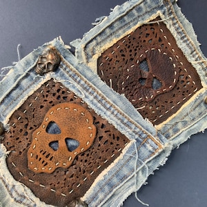 Könnte beinhalten: Zwei quadratische Jeans-Aufnäher mit ausgefransten Kanten und dunkelbraunen Lederauflagen. Jedes Lederstück weist ein lasergeschnittenes Filigranmuster und einen mittigen Totenkopf-Ausschnitt auf, befestigt mit weißen Nähten.