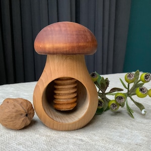 Könnte beinhalten: Ein pilzförmiger Nussknacker aus Holz mit brauner Kappe und hellem Holzkörper. Eine Walnuss liegt neben dem Nussknacker. Dekorative grüne und gelbe Beeren befinden sich im Hintergrund.