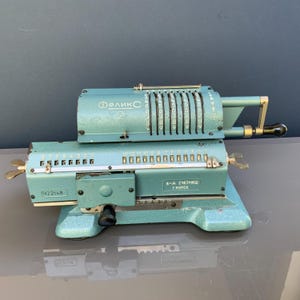 Op de afbeelding: Een vintage turquoise mechanische rekenmachine met de naam "Феликс" en het nummer "2" erop gedrukt. De rekenmachine heeft meerdere nummerwielen en een slinger. De tekst "3-А СЧЕТМАШ Г КУРСК" is ook zichtbaar.