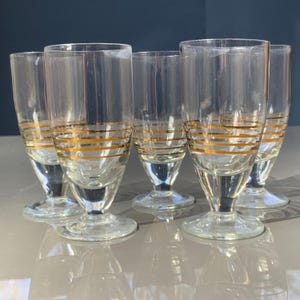Op de afbeelding: Set van zes heldere likeurglazen met gouden horizontale strepen. De glazen hebben een uitlopende bovenkant, een korte steel en een ronde voet. De gouden strepen voegen een vleugje elegantie toe aan het vintage glaswerk.