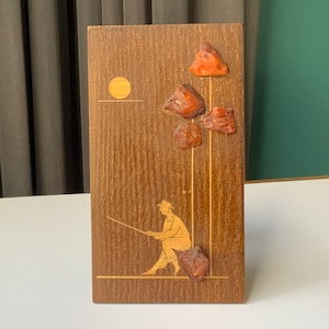 Peut inclure: Plaque en bois représentant une scène de pêche. L'œuvre d'art comprend un pêcheur, un soleil et des éléments décoratifs. La plaque a un grain de bois naturel et est ornée de trois accents texturés brun-rouge.