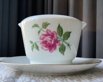 May include: A white porcelain gravy boat with a pink rose and green leaves design. The boat has a matching saucer with a gold trim.