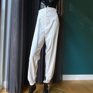 May include: Cream-colored high-waisted pants with a button closure and a tapered leg. The pants are displayed on a mannequin, showcasing their tailored fit and design. The fabric appears to be lightweight, suitable for warmer weather.