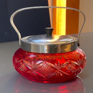 Puede incluir: Un azucarero de vidrio rojo con tapa y asa de plata. El cuenco tiene un diseño de vidrio cortado.